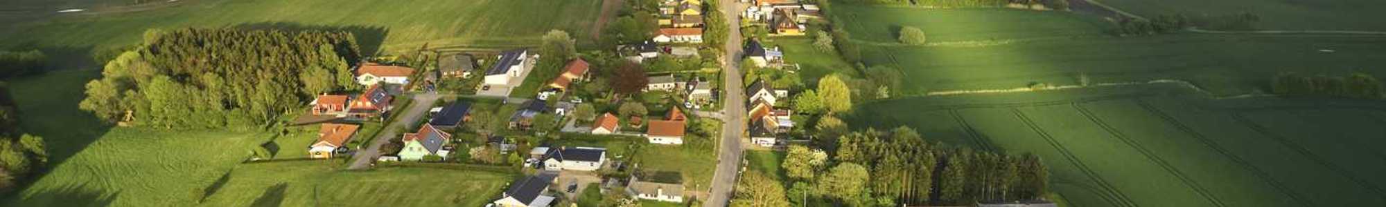 Luftfoto af en lille landsby omgivet af grønne marker og vindmøller i det fjerne.